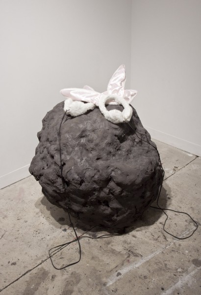 A sculpture made of dark organic material with rounded shapes and two pairs of headphone sit fur and pink bunny ears