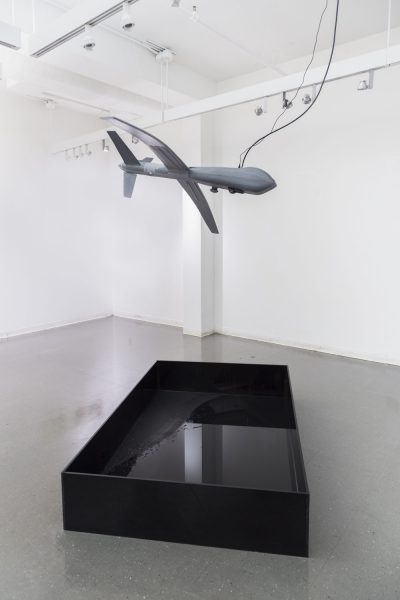 Grey airplane sculpture hanging from the ceiling above a black box on the ground filled with a shallow amount of water that looks reflective.