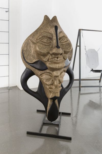 Sculpture of a large stone-like mask on top of a black stone-like face on a black wooden stand.