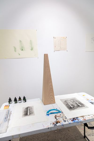 Artwork by Marc Cioffi. A scale model of a wooden sculpture on top of a table, prints of pyramid shape objects.