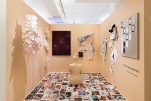 Installation view of artwork by Maria Barquet. Two mixed media paintings, Cloth attached with thread attached to the wall, two sculptures made of cut cloth squares that are suspended from the ceiling, various torn pages from magazines arranged in a grid covering the floor and large flesh toned sculpture of a woman sitting meditating toward the rear of the space.