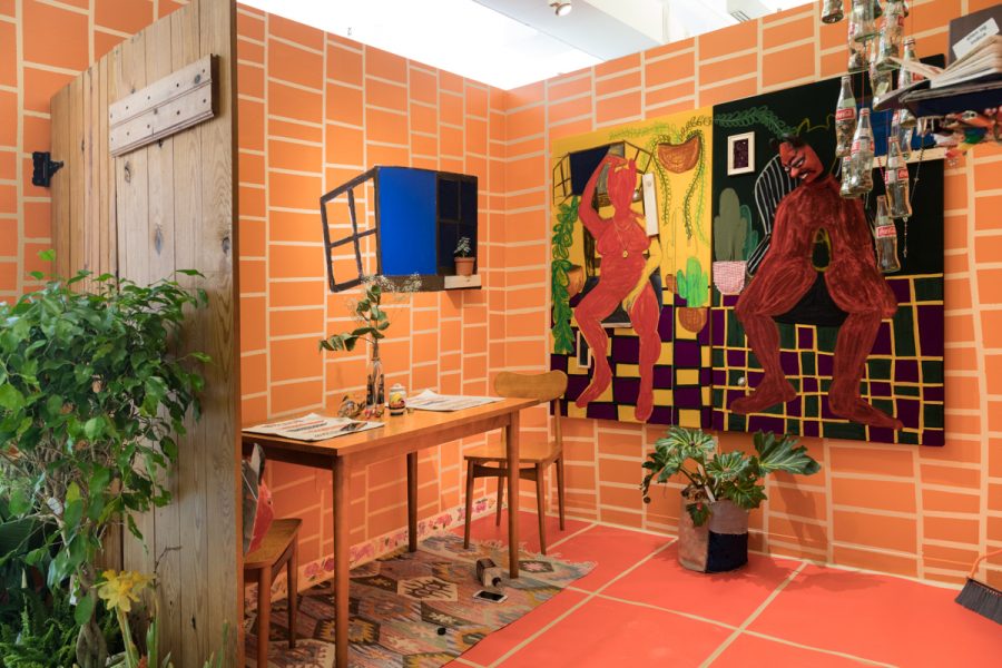 Installation view of artwork by Maria Barquet. Fabricated dinning nook with faux brick walls, wooden fence, potted plants and two abstract paintings of figures using warm hues.