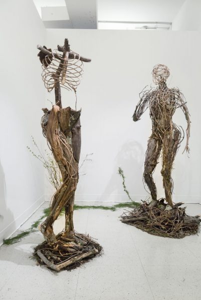 Two sculptures of the human form in various poses, made from sticks, twigs, and ivy, on a piles of dirt.