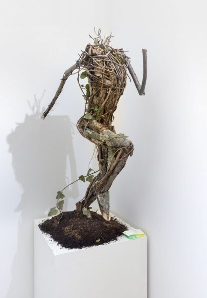 Sculpture of the human form in a running pose made from sticks, twigs, and ivy, on a pile of dirt on a pedestal.