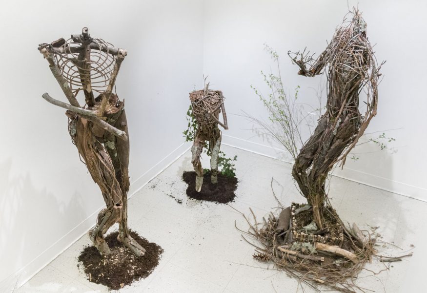 Three sculptures in the corner of a room made from twigs, sticks, and natural wood, of the human form.