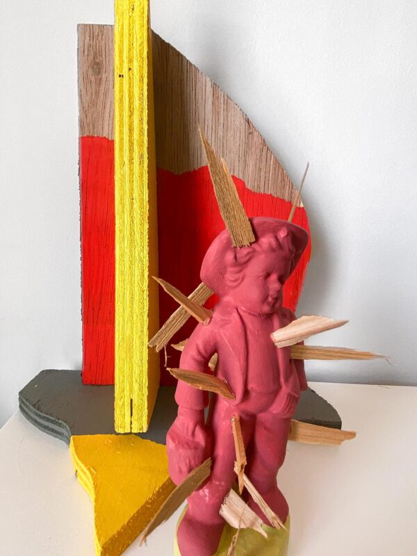 Detail shot of small sculpture, a red baby stabbed with wooden splinters against a wooden abstract background.