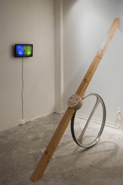 Installation view of a sculpture made with a medium steer wheel with a transversal bar inside the wheel shape, a long wood piece fixed diagonally on the floor, a portion sitting on the wheel, and the top part is on the wall, in the back is a TV with an image displayed with blue and green light