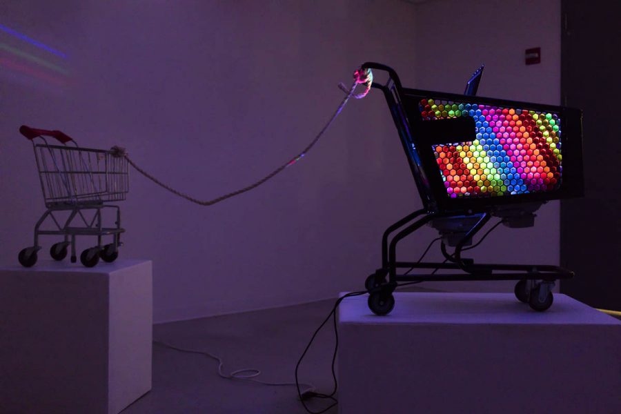 In a dark room, there is a shopping cart installed on a pedestal with RGB lights inside the cart, and behind it is linked with a wire, a smaller but empty cart