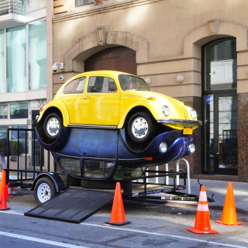 Angle view of two Volkswagen Beetles, a yellow one on top and a blue one at the bottom, welded together from the underside of each car, sitting on a towing trailer. The Blue one is at the bottom, sitting on its rooftop, and the yellow is on top of the blue car.