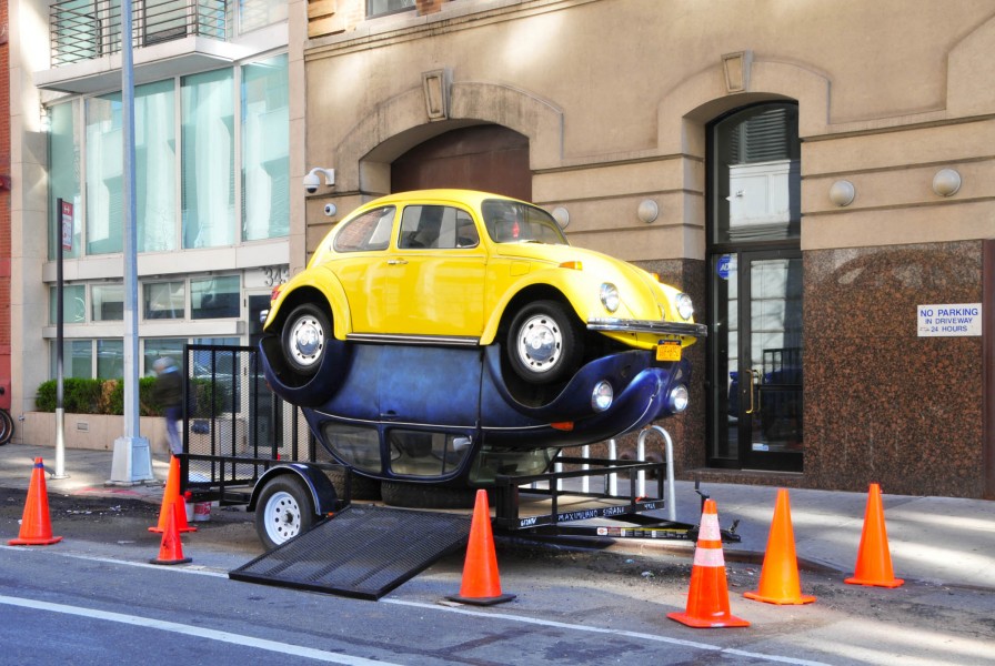 Angle view of two Volkswagen Beetles, a yellow one on top and a blue one at the bottom, welded together from the underside of each car, sitting on a towing trailer. The Blue one is at the bottom, sitting on its rooftop, and the yellow is on top of the blue car.