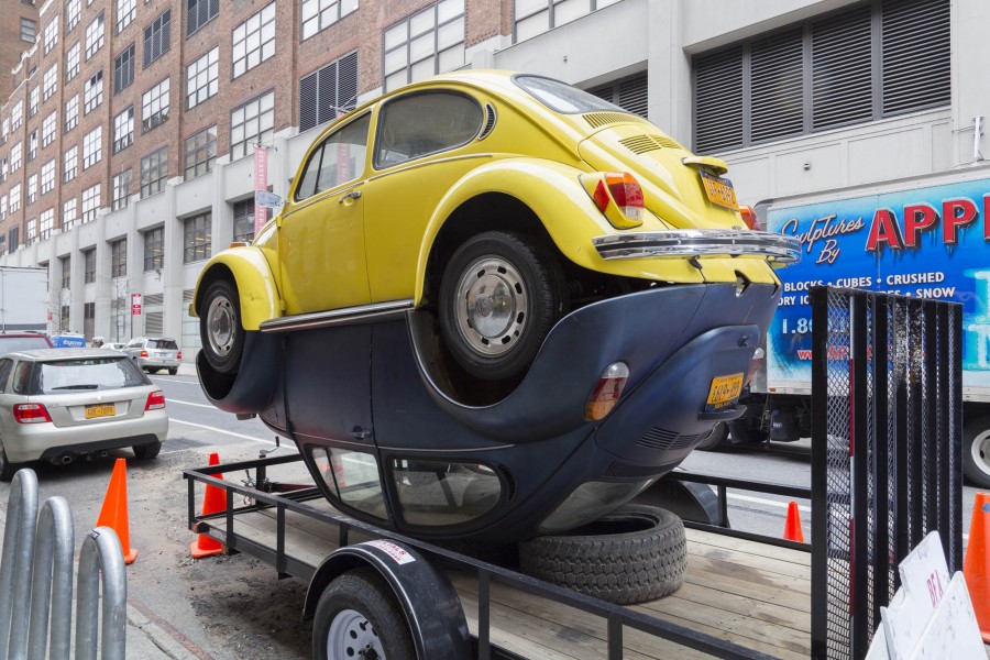 Back-view of two Volkswagen Beetles, a yellow and a blue one, welded together by the underside of the cars like one car is sitting on the rooftop on the ground and one is sitting in a normal position. The cars are sitting on a towing trailer in the street.