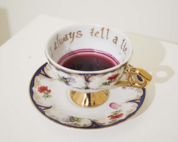 Teacup win golden handle and some tea on the bottom of the cup, a written gold message saying I always tell a lie, the cup is put on a small plate with blue, red, and golden motives