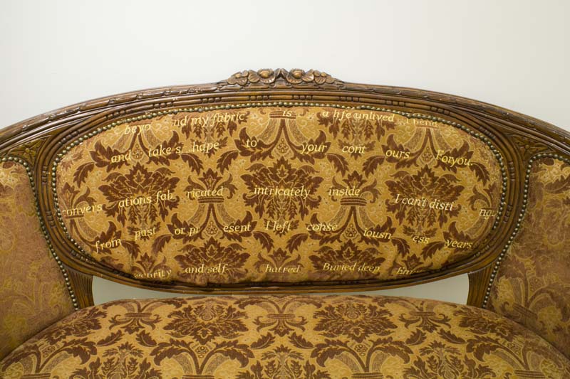 A vintage golden couch made of wood and fabric material with vintage patterns and a text with generously spaced words on the backrests