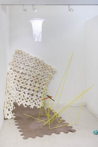 Installation view of a sculpture composed from a brown ridged matt on the floor, a yellow angular structure made with straight bars, a big matt which looks to be made out of pieces of toilet paper near a wall, and a light with an undershirt as a lampshade