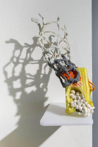  A small sculpture installed on a white shelf made with organic yellow, orange, grey and white materials, with rounded shapes, the main body has a rectangular shape with an opening and some small white balls in it, and a mesh-like formation goes up on the left corner