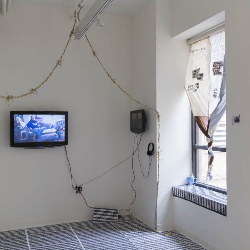 Installation view of two TVs installed each on a separate wall and a window covered partially with three pieces of curtains sewed together and with text on the, twisted at the base of the window