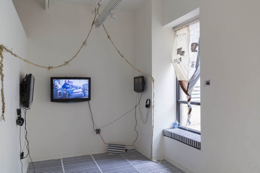 Installation view of two TVs installed each on a separate wall and a window covered partially with three pieces of curtains sewed together and with text on the, twisted at the base of the window
