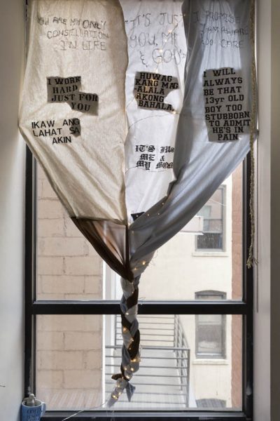 A view of a window with three window curtains sewed together with text on them, and at the base of the window, they are twisted together and not covering the window