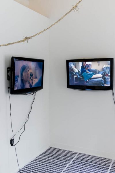 Installation view of two tv screens displaying an image with a girl looking and talking to a guy, and the other tv is displaying an image with an older woman brushing the hair of a girl in the living room