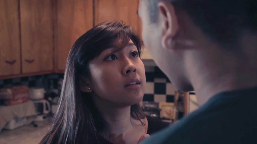 A still image from a movie with a girl facing up to talk to a man, in a kitchen with cabinets installed on the wall and different appliances