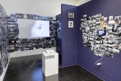 Installation view of dozens of photographs installed on walls, a video projector, and its projection in the wall in the background