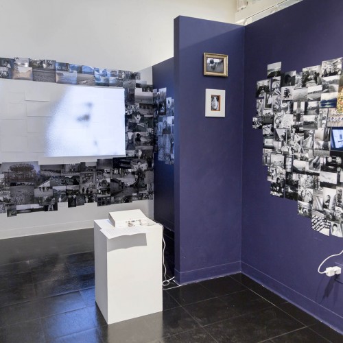 Installation view of dozens of photographs installed on walls, a video projector, and its projection in the wall in the background