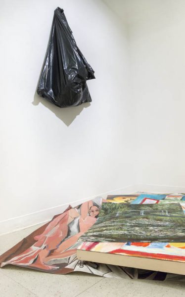 Installation view of a pile of paintings with portraits and landscapes on the floor, and a black garbage bag hung on the wall with something in it