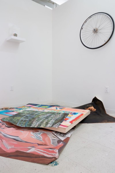 Installation view of paintings piled up on the floor with the landscape, room interior in front of the windows, a nude painting of a woman, a portrait of a man, a shelf with a Starbucks coffee cup on a transparent container, and a bike wheel suspended on the wall