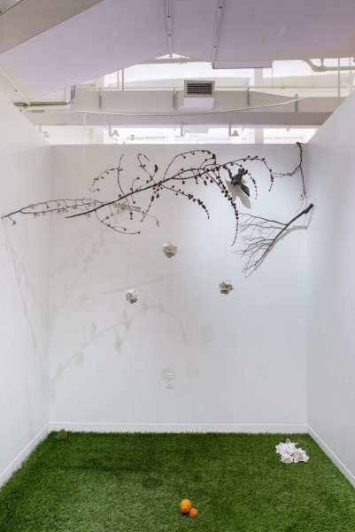 Installation view of tree branches suspended in the air, a stuffed bird, three white floral objects are hanging from the branches, fake grass on the floor with many objects on it.