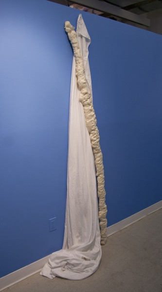 A thin vertical sculpture made of organic material with rounded and bubbly shapes and a white cloth hung on the blue wall.