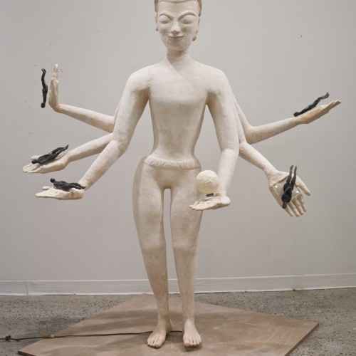 Front view of a white, anthropomorphic sculpture with six hands with palms wide open, and one palm has a rounded ball in it, but the others have a small black anthropomorphic sculpture