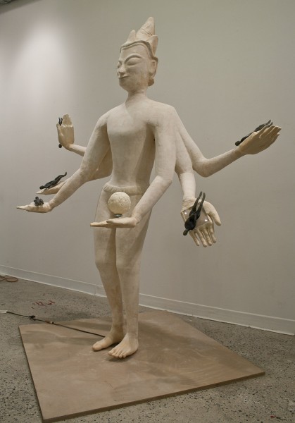 Angle view of a white, anthropomorphic sculpture with six hands with palms wide open, and one palm has a rounded ball in it, but the others have a small black anthropomorphic sculpture