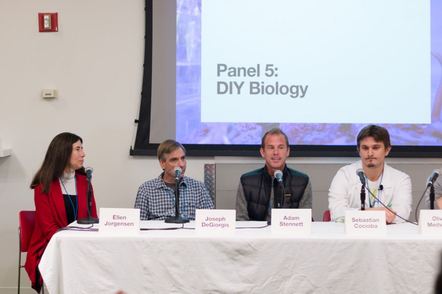 Ellen Jorgensen, Joseph DeGiorgis, Adam Stennett, and Sebastian Cocioba are sitting on the desk, and behind them is a projection of Panel 5: DIY Biology