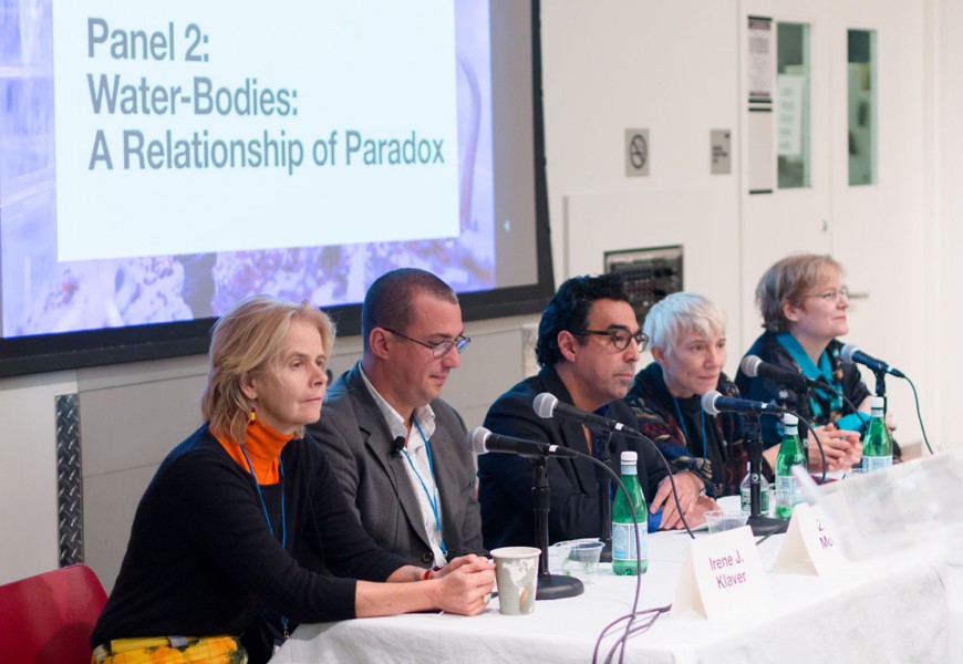 Alumni members sitting at the table with microphones and talking to the public; behind them is a projection of Panel2: Water-Bodies: A Relationship of Paradox