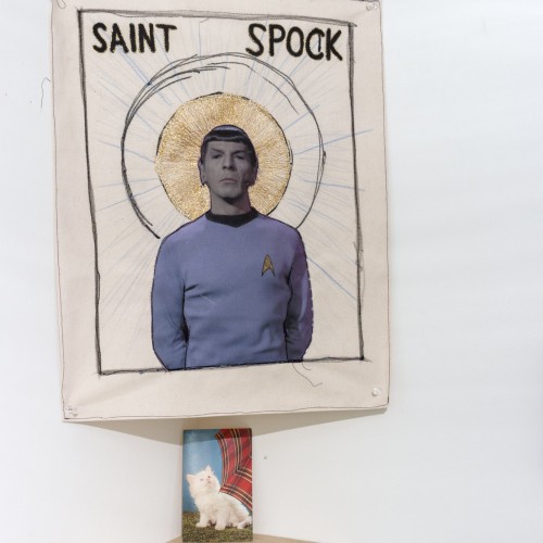 Composite work made of a cut-out of Spock from Star Wars with a saint aura and SAINT SPOCK written at the top, two small prints of a kitten, and a led candle light on a shelf.