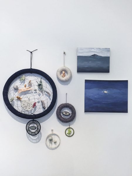 Five dream catchers are installed on a white wall, the one to the left is the biggest and the other four dream catchers are very small, they are navy blue or white, with eye motifs and other various objects woven in the middle, to the right are two rectangular paintings of the ocean, they are gray, blue, and white
