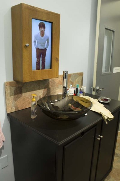 Installation view of bathroom shelf with door and an image of a man with hand in the pocket on it, a sink installed on black bathroom furniture, and different objects