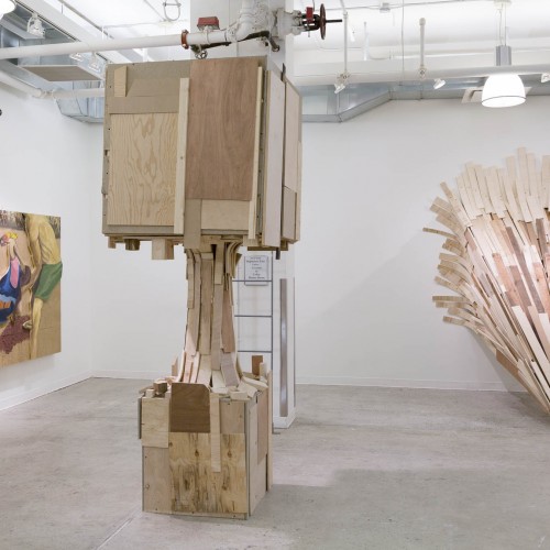 Installation view of two wooden sculptures, one conic-shaped in the right of the room, in the middle of the room is one sculpture with a small cube as the base, a pillar and a bigger cube, double-sized of the base at the top, on the left side of the room is an oil painting made on a sanded wood with pieces cut in different shapes illustrating sexual aggression of a woman.