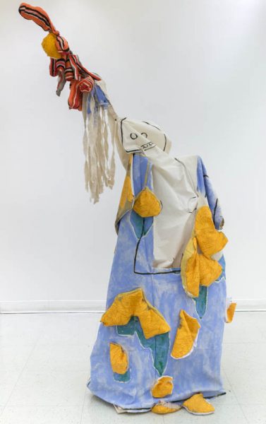 A sculpture made of organic fabric painted in blue and lots of yellow pockets. It has the shape of a human body with a drawing of a face on the head, and it had a hand raised with a piece of fabric with red, black, and yellow stripes