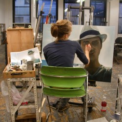 A student works on a portrait with a man wearing a white hat, she is in front of an easel on a chair, and she has a crate with painting materials and a reference photo to guide her painting