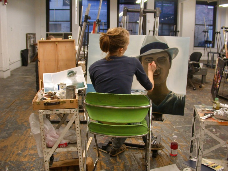 A student works on a portrait with a man wearing a white hat, she is in front of an easel on a chair, and she has a crate with painting materials and a reference photo to guide her painting