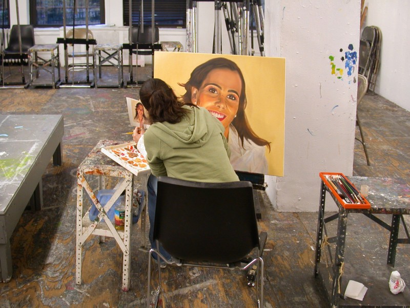 A student is working on a portrait of a woman with a big smile on a yellow background. The student has brushes and paints on a working table near her.