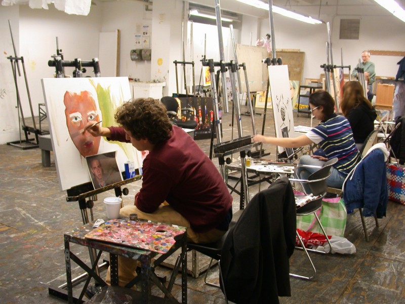 A student works on a portrait with a brush in hand, paint near him, in the background, other students work on paintings on easels