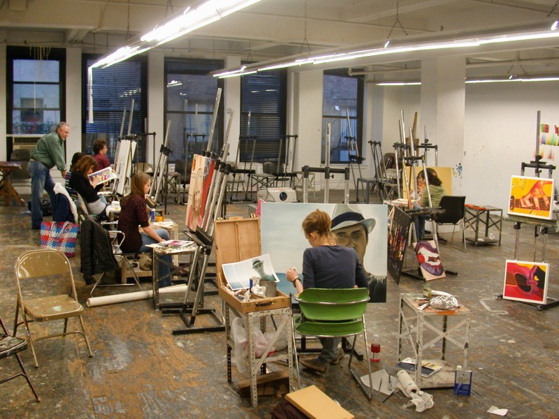 Painting class with students working on portraits, sitting on chairs with easels in front of them, paintings are laying around in the class on supports, working materials such as paint, brushes, etc.