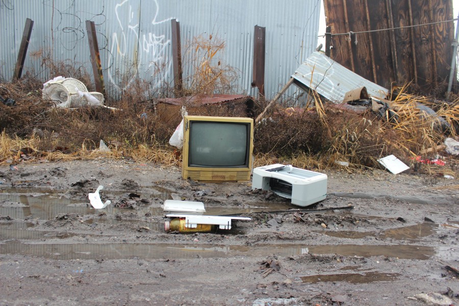 There is a landscape of an old yellow TV placed on a muddy road, a white printer on the right side of the TV, other objects thrown on the ground, and a metal fence in the background.