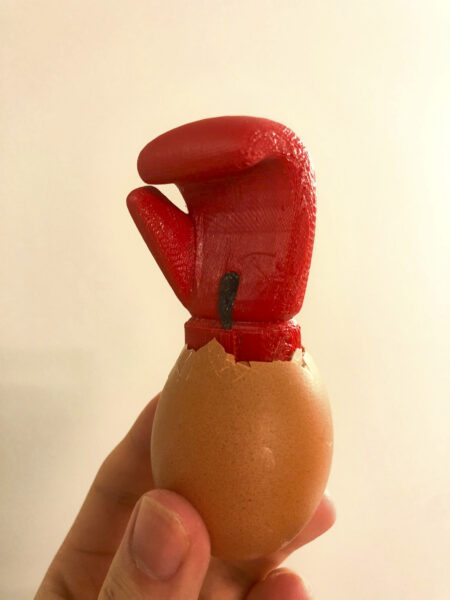 A 3D-printed red boxing glove emerging from the shell of a hen's egg, which is held by a human hand.