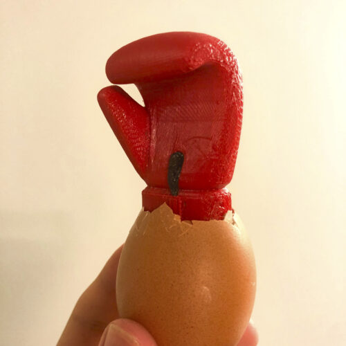 A 3D-printed red boxing glove emerging from the shell of a hen's egg, which is held by a human hand.