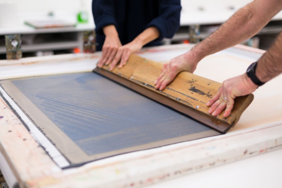 A large squeegee being pulled across a large silkscreen frame. An image of landscape can be seen on the frame.