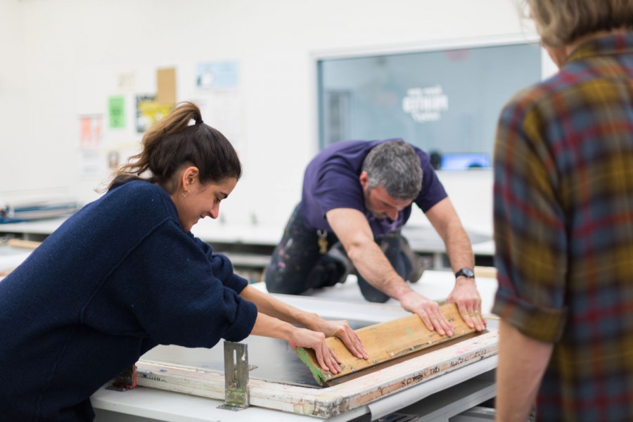 An instructor helps a student pull a large squeegee across a silkscreen frame.