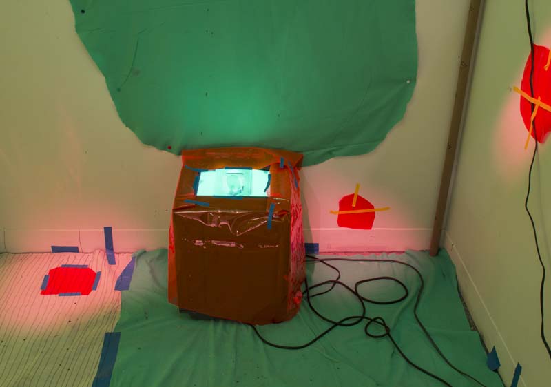 Small screen installed in a box covered with red transparent foil, green fabric installed on the wall and the floor, and small red fabrics installed on the wall and floor with red spotlights hitting them.